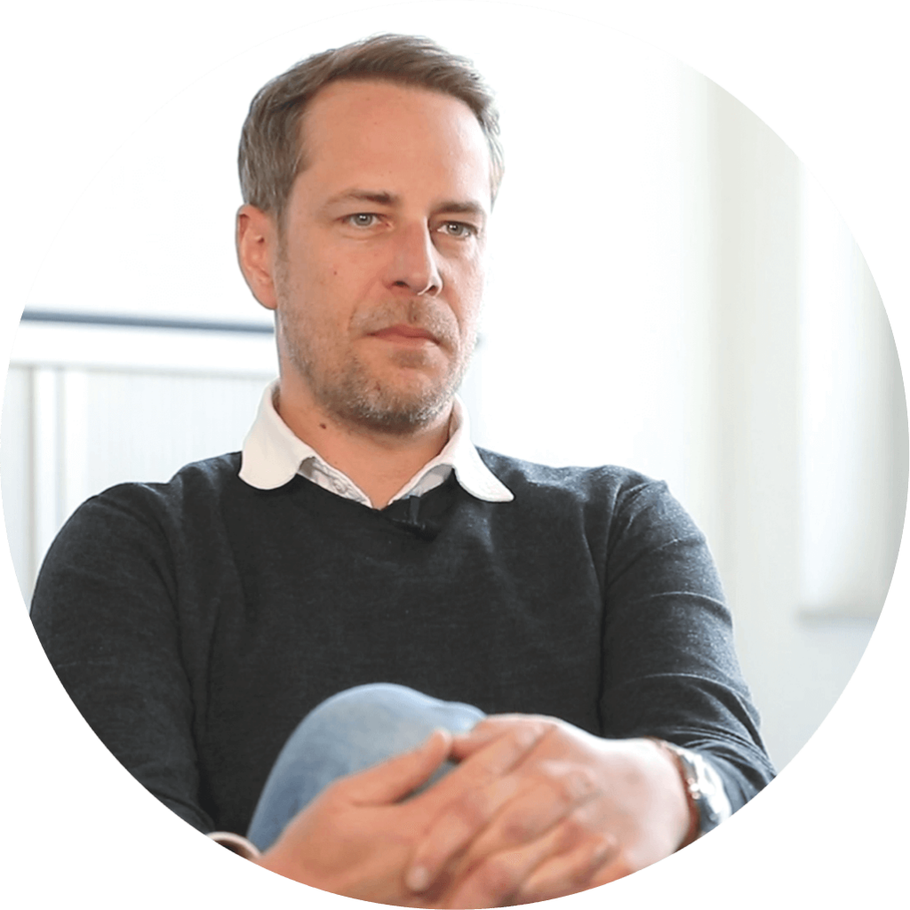 Porträt eines erwachsenen Mannes mit verschränkten Armen in ruhiger Sitzposition. Er trägt einen dunklen Pullover über einem weißen Hemd und blickt konzentriert nach vorne. Professionelle Business-Porträtaufnahme in hellem Innenraum, geeignet für Unternehmensdarstellung, Beratung, Management- oder Expertenprofil im Pflege- und Gesundheitsumfeld.